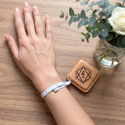 Hand with a measuring tape, tan leather case with logo, and white rose on wooden surface